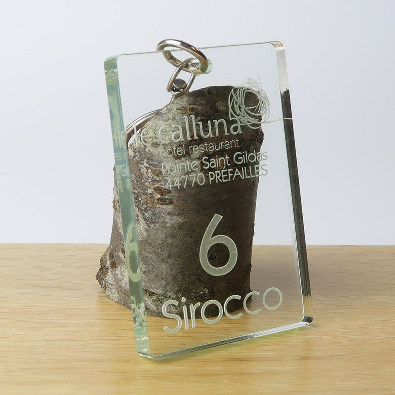 Porte-clé de 9 cm x 6 cm x 0,8 cm en plexiglass transparent, gravé avec le logo de l’hôtel Calluna, le numéro de chambre, et le nom de la chambre sur la face avant.